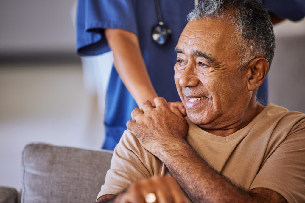 Caregiver in scrubs with her hand on an elderly man's shoulder who sits in front of her.